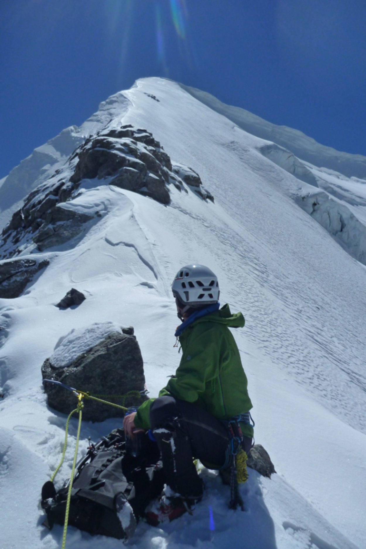 Hochtourengeher sitzt am Gipfelgrat, sichert mit einem Seil den Nachgeher und schaut in Richtung Gipfel des Piz Palü.