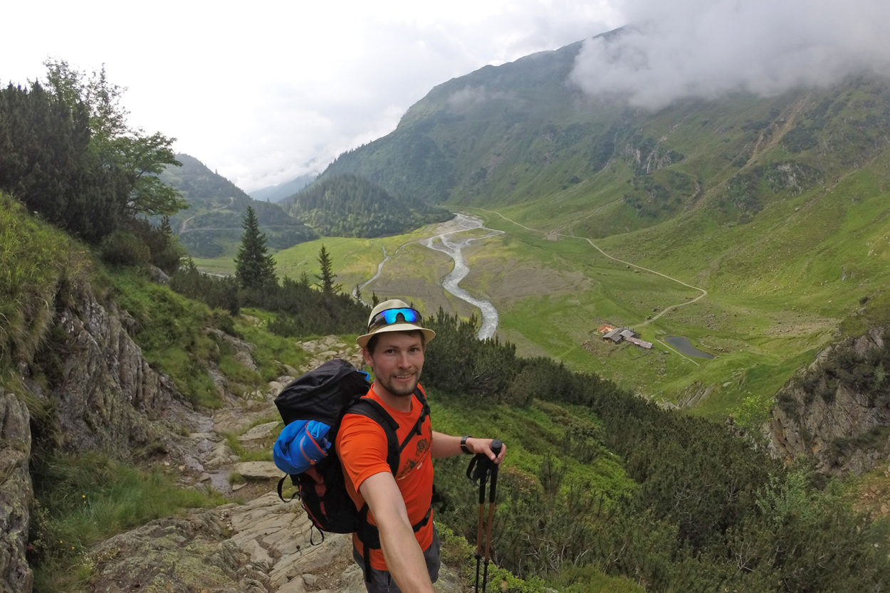 Ein Bergsteiger macht ein Selfie mit Blick auf eine grüne Hochalm, durch die sich ein Weg zieht.