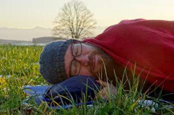 Ein Mann liegt auf einer Wiese mit dem Kopf auf der verpackten Icebreaker Hyperia Lite Hybrid Jacke.